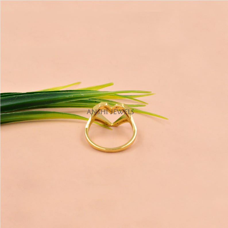 May include: A gold-colored ring in the shape of a heart, with the brand name "ANSHI JEWELS" visible. The ring is set against a soft pink background, with green leaves in the upper left corner.