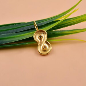 May include: A gold-colored pendant in the shape of an infinity symbol, with two small gemstones set within the loops. The pendant has a loop at the top for a chain. The background is a soft peach color with green leaves.