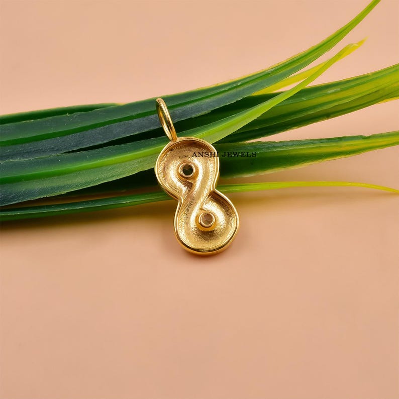 May include: A gold-colored pendant in the shape of an infinity symbol, with two small gemstones set within the loops. The pendant has a loop at the top for a chain. The background is a soft peach color with green leaves.