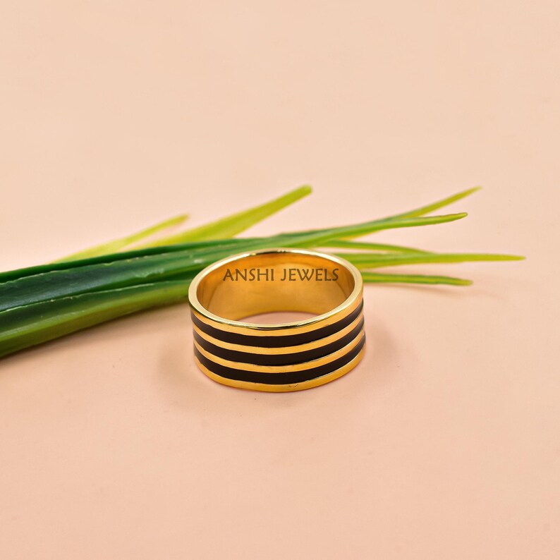 May include: A gold-colored ring with three black enamel stripes. The ring is set against a soft pink background with green leaves. The text "ANSHI JEWELS" is visible on the ring.