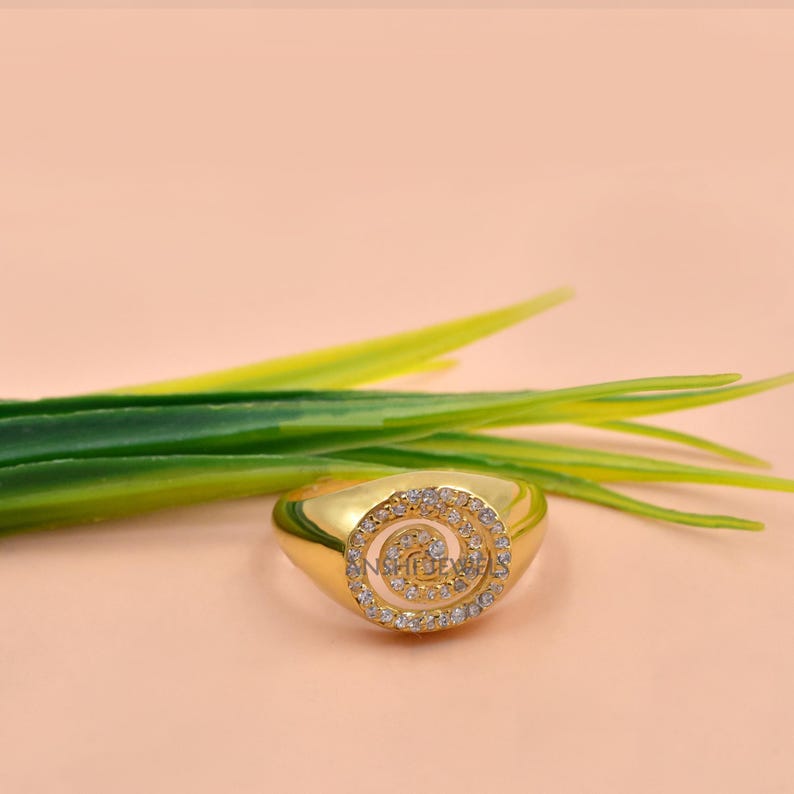Puede incluir: Un anillo de tono dorado que presenta una cara circular con un patr&oacute;n en espiral incrustado con peque&ntilde;as piedras claras y brillantes. La banda es lisa y pulida, mostrando un acabado met&aacute;lico continuo que complementa el dise&ntilde;o central.