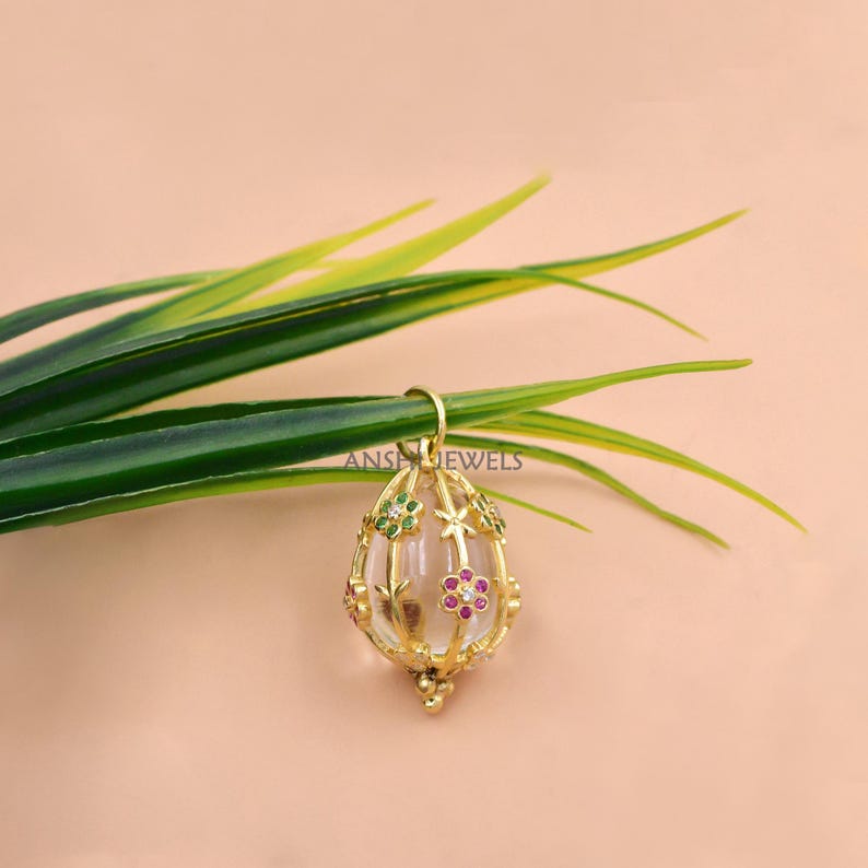 Pode incluir: Pingente dourado com um centro transparente em forma oval. O pingente &eacute; adornado com pequenas pedras preciosas coloridas e desenhos florais. O texto "ANS JEWELS" &eacute; vis&iacute;vel no la&ccedil;o. O pingente est&aacute; pendurado em uma folha verde.