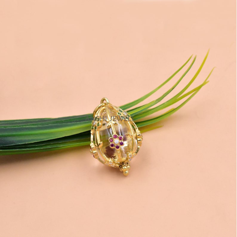 Pode incluir: Um pingente dourado com um centro transparente em forma de ovo, adornado com uma pequena flor vermelha e detalhes brancos. O pingente tem intrincados detalhes em filigrana dourada e uma pequena base decorativa. O fundo &eacute; de cor p&ecirc;ssego suave com uma folha verde.
