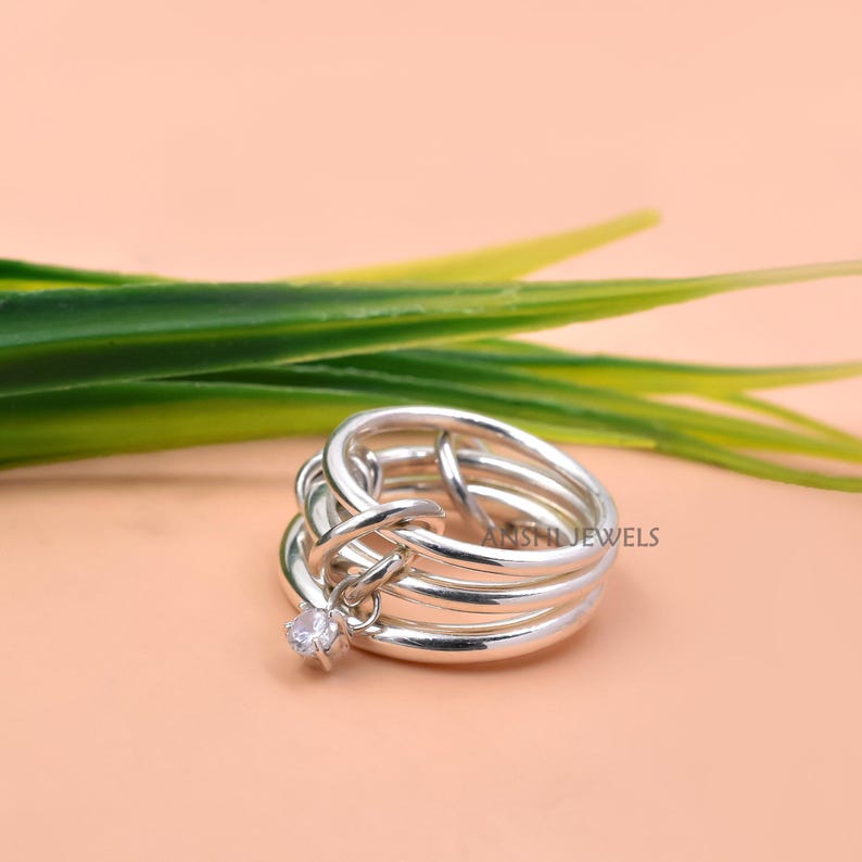 May include: A silver-toned multi-band ring featuring a polished finish and an interlocking design. A single small, clear, round-cut gemstone is suspended from one of the bands as a charm. The text ANSHIJEWELS is visible on the metal surface.