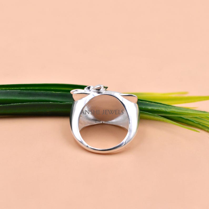 May include: A silver-toned ring featuring a smooth, rounded band and a decorative top element. The interior of the ring band is clearly engraved with the text ANSHI JEWELS in a clean, sans-serif font, visible through the open center of the ring.