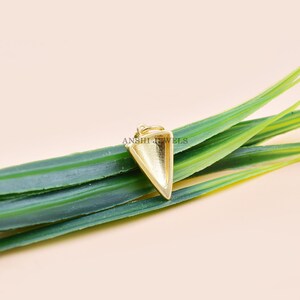 Pu&ograve; includere: Un pendente dorato a forma triangolare con superficie strutturata, sospeso a un piccolo anello. Poggia su una foglia verde su uno sfondo color pesca chiaro. Il testo "ANSHI JEWELS" &egrave; visibile. Un gioiello.