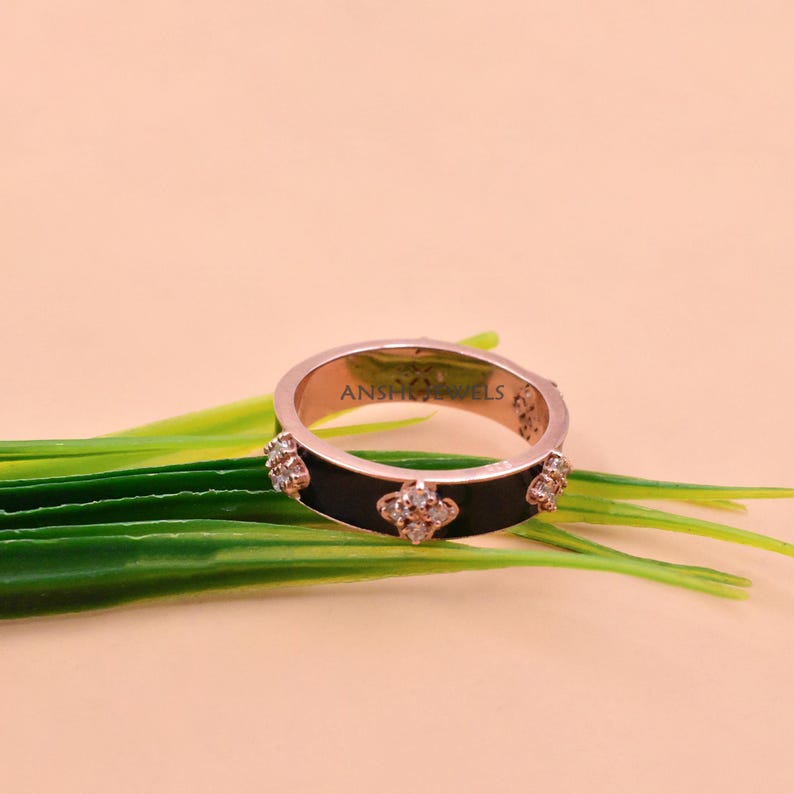 May include: A rose gold band ring featuring a black enamel inlay and repeating four-petaled flower motifs set with small clear stones. The interior of the band is engraved with the text ANSH JEWELS. The ring has a polished metallic finish.