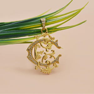 May include: A gold-colored pendant in the shape of the letter "C", adorned with small, sparkling stones. The pendant features an ornate, floral design with delicate curves and embellishments. The pendant is suspended from a small loop, and the brand name "ANSHI JEWELS" is visible.