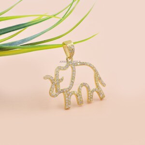Op de afbeelding: Een gouden olifant hanger met heldere zirkonia stenen. De hanger is op een licht roze achtergrond.