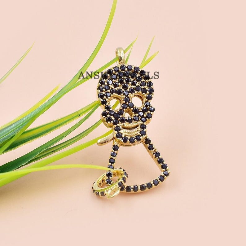 Puede incluir: Pendiente de tono dorado con un dise&ntilde;o de calavera adornado con piedras de circonita c&uacute;bica negras. La calavera lleva un anillo en el dedo.