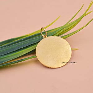 May include: A gold-colored, round pendant with a small loop at the top. The pendant has a textured surface and the text "ANSHI JEWELS" is visible. The pendant is set against a backdrop of green and yellow leaves and a pink background.