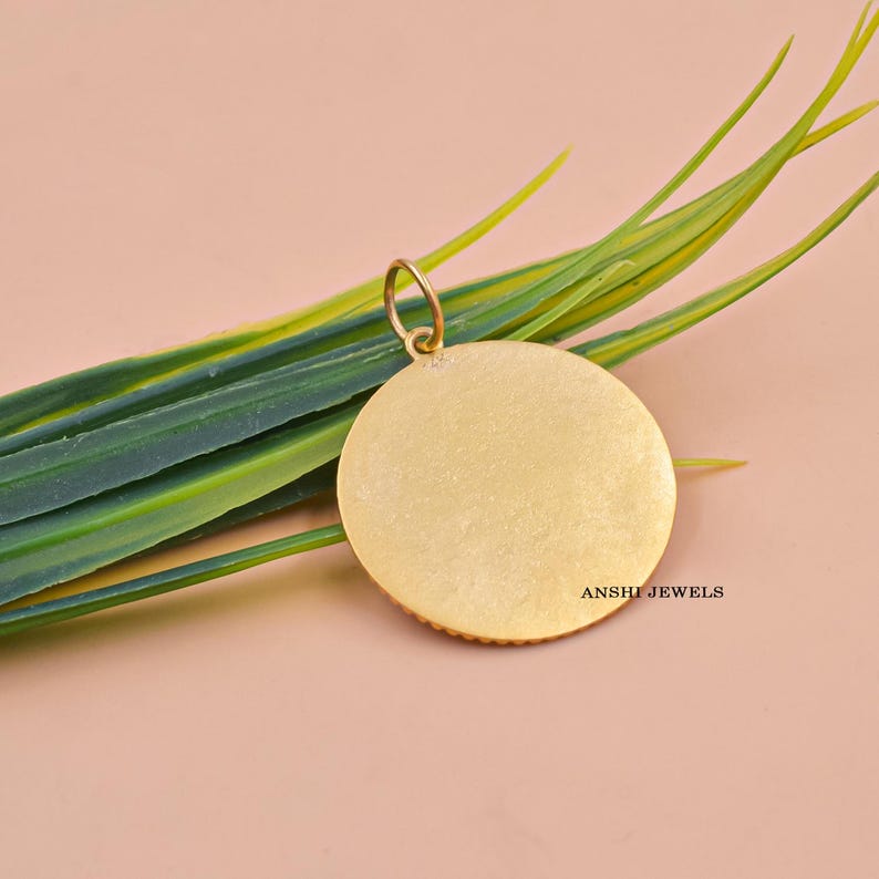 May include: A gold-colored, round pendant with a small loop at the top. The pendant has a textured surface and the text "ANSHI JEWELS" is visible. The pendant is set against a backdrop of green and yellow leaves and a pink background.