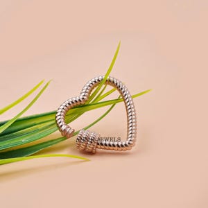 K&ouml;nnte beinhalten: Ein herzf&ouml;rmiger Verschluss in Ros&eacute;gold mit einem strukturierten, seilartigen Design. Der Verschluss ist ge&ouml;ffnet und gibt den Mechanismus frei. Der Hintergrund ist ein weicher Pfirsichton mit gr&uuml;nen Bl&auml;ttern.