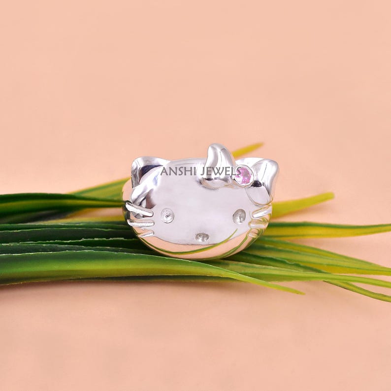 May include: A silver-toned ring shaped like a cat face with whiskers, featuring a small pink stone set in the bow and clear stones for the eyes and nose. The text ANSHI JEWELS is engraved on the forehead of the cat-shaped ring design.