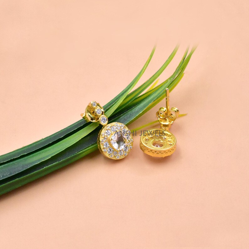 Op de afbeelding: Goudkleurige oorbellen met een ronde, heldere edelsteen in het midden, omringd door kleinere heldere stenen. De oorbellen zijn geplaatst op een lichtroze achtergrond met groene bladeren. De tekst "ANSHI JEWELS" is zichtbaar.