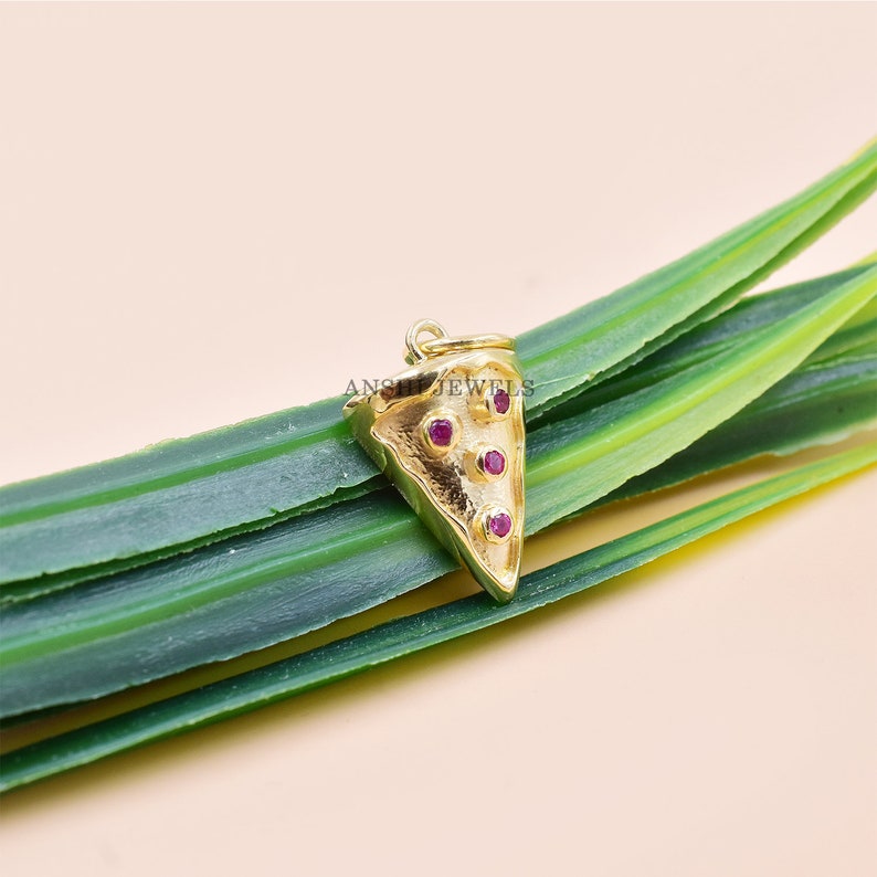 Pu&ograve; includere: Un ciondolo a forma di fetta di pizza color oro con tre piccole gemme rotonde e rosse. Il ciondolo &egrave; sospeso da un piccolo anello nella parte superiore. Il ciondolo &egrave; appoggiato su una foglia verde.