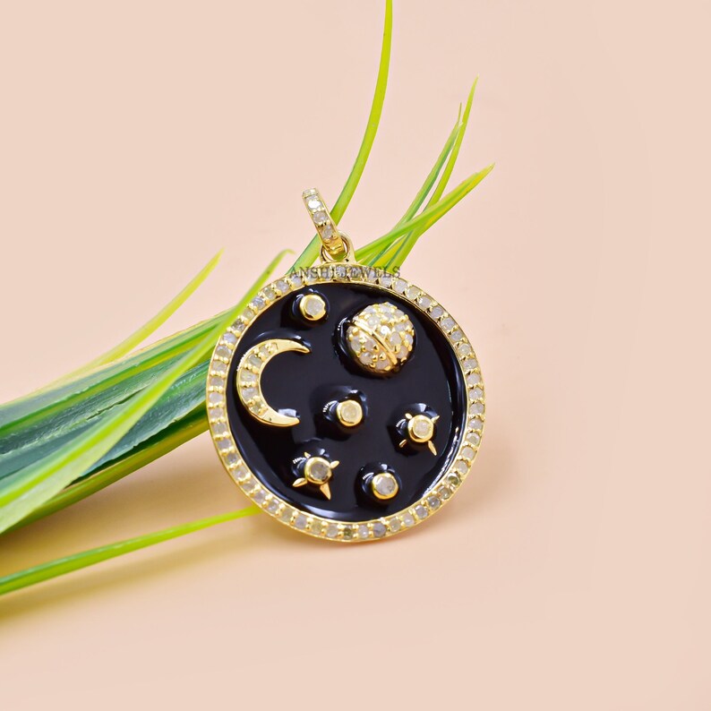 May include: Gold pendant necklace with a black enamel circle featuring a crescent moon, a planet, and stars. The circle is surrounded by a border of small clear stones.