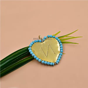 May include: A heart-shaped pendant with a gold-colored surface and a turquoise bead border. The pendant has the letter "W" cut out of the gold. The pendant is on a green leaf.