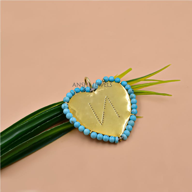 May include: A heart-shaped pendant with a gold-colored surface and a turquoise bead border. The pendant has the letter "W" cut out of the gold. The pendant is on a green leaf.
