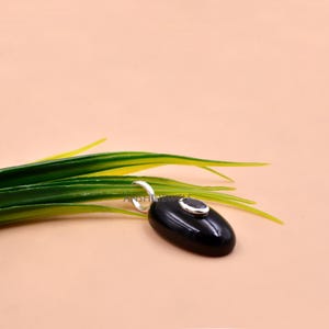 May include: An oval-shaped black onyx pendant featuring a small, centered, oval-cut gemstone set in a silver bezel. The pendant includes a silver bail at the top for attachment. The piece is positioned against a light peach-colored background.