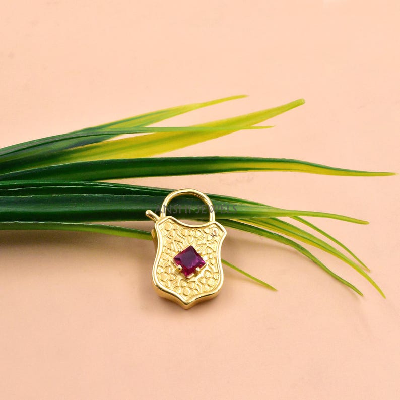 May include: A gold-colored shield-shaped pendant with a square, deep red gemstone at its center. The pendant has a floral pattern and a small loop at the top. The background is a light peach color with green and yellow leaves.