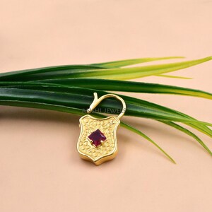 May include: A gold-colored shield-shaped pendant with a square, deep red gemstone at its center. The pendant has an ornate, textured design and a curved bail. The background includes green and yellow leaves.