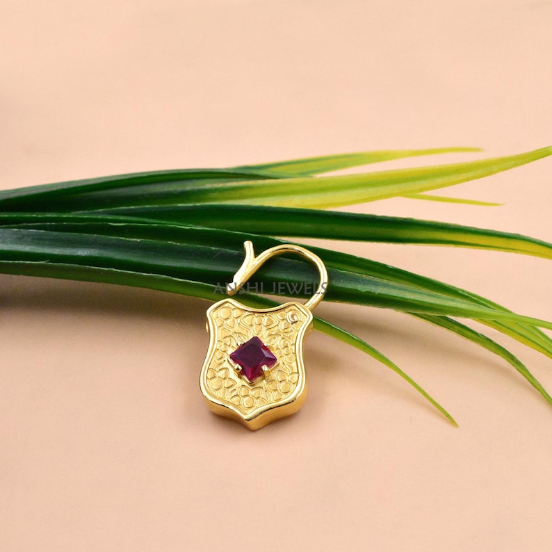 May include: A gold-colored shield-shaped pendant with a square, deep red gemstone at its center. The pendant has an ornate, textured design and a curved bail. The background includes green and yellow leaves.