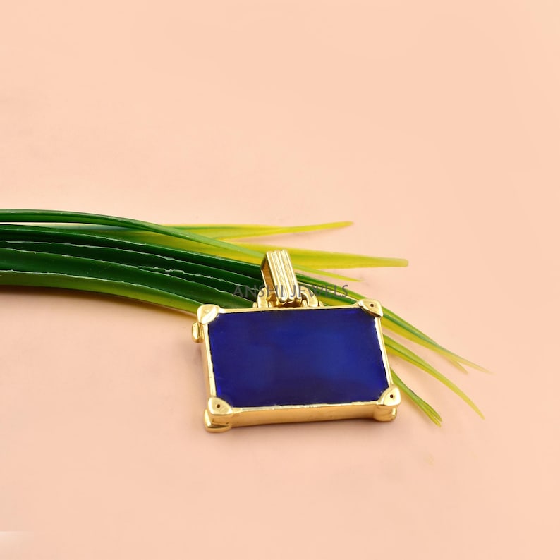 May include: A rectangular pendant featuring a deep blue center inlay framed by a polished gold-tone metal border. The design includes four small rounded corner accents and a vertical grooved bail at the top for attachment to a necklace chain.
