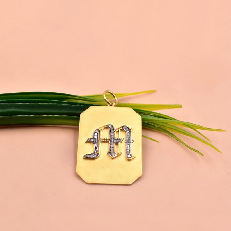 May include: A rectangular gold pendant featuring a gothic-style letter M encrusted with small clear stones. The pendant has a smooth matte finish and a small circular bail at the top for attachment. The text AISHI JEWELS is visible on the letter.