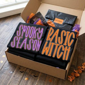 May include: Two black t-shirts folded inside a cardboard box. One shirt features the purple text SPOOKY SEASON, and the other features the orange text BASIC WITCH. A black envelope with a boutique label is tucked behind the shirts with tissue paper.