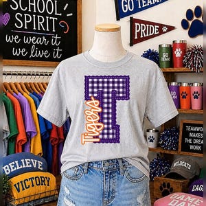 May include: A light gray short-sleeve t-shirt featuring a large purple and white gingham patterned letter T with a decorative stitched border. The word Tigers is printed in orange cursive script vertically along the left side of the letter T.