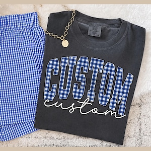 May include: A charcoal gray t-shirt featuring the word CUSTOM in a blue and white gingham block font, with the word custom written below in white script. Beside the shirt are blue and white gingham shorts and a gold-toned chain necklace.