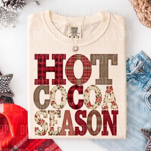 May include: A cream-colored short-sleeve t-shirt featuring the text HOT COCOA SEASON in large block letters. The letters are filled with various patterns including red plaid, brown burlap, and a cream floral print. The shirt has a Comfort Colors label.