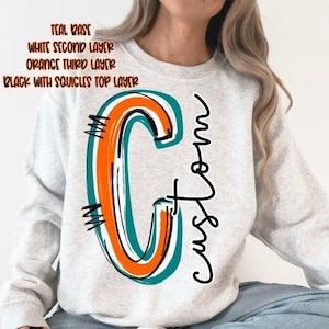May include: A light gray crewneck sweatshirt featuring a large graphic of the letter C layered in teal, white, orange, and black with squiggles. The text "custom" is written in black cursive script next to the letter. The image includes text labels.