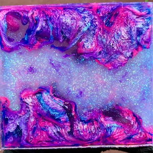 May include: Square resin artwork featuring a swirling design in purple, pink, and blue hues. The piece has a glittery, galaxy-like aesthetic, with a smooth, glossy finish. The colours blend to create an abstract, ethereal effect, reminiscent of a nebula.