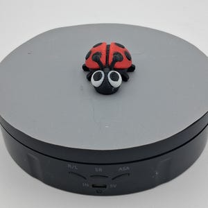 May include: A small, red and black ladybug figurine with large white eyes and black spots. The ladybug is resting on a gray, circular platform. The platform sits on a black base with control buttons.