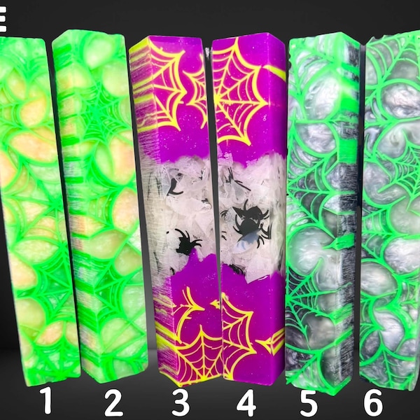 May include: Six rectangular resin pen blanks labeled E, arranged in pairs. The first pair is neon green with yellow swirls and spiderweb patterns. The second pair is purple with yellow spiderwebs and embedded black spider shapes. The third pair is black and white with neon green spiderweb patterns.