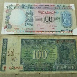 May include: Two Indian 100 Rupee banknotes. The top note is mainly white and pink, with text in multiple languages and the Reserve Bank of India seal. The bottom note is green and tan, also featuring text in multiple languages and the Reserve Bank of India seal.