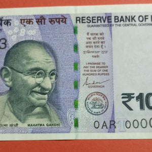 Könnte beinhalten: Ein einhundert indische Rupien-Banknote. Die Banknote zeigt ein Porträt von Mahatma Gandhi, den Text "RESERVE BANK OF INDIA" und die Zahl "100". Die Banknote ist hauptsächlich lila und weiß.