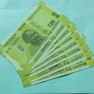 May include: A fan of Indian twenty rupee banknotes, featuring a portrait of Mahatma Gandhi. The notes are primarily green with the denomination clearly visible. Each bill displays a unique serial number.