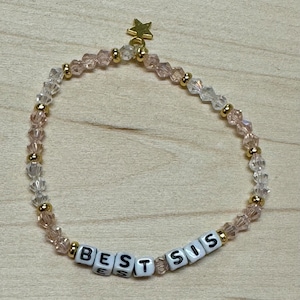 May include: A delicate bracelet with a gold star charm and clear and pink crystal beads. The bracelet has the word "BEST SIS" spelled out in white letter beads.