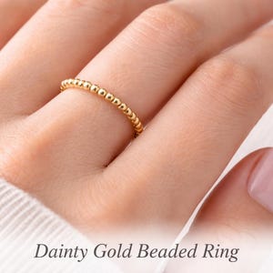 Puede incluir: Un delicado anillo de cuentas doradas. El anillo presenta una línea continua de pequeñas cuentas espaciadas uniformemente. El anillo se muestra en un dedo, con el texto "Dainty Gold Beaded Ring" en la parte inferior.