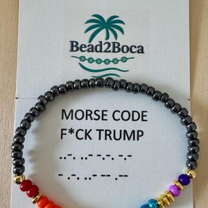 May include: A beaded bracelet with a rainbow gradient and gold accents. The bracelet features the text "MORSE CODE F*CK TRUMP" on a white card. The bracelet is made of small, dark gray beads.