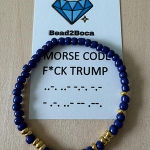 May include: A beaded bracelet with navy blue beads and gold-colored spacers. A card behind the bracelet reads "MORSE CODE F*CK TRUMP" with the Morse code translation. The brand name "Bead2Boca" is also visible.