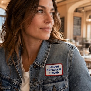 Puede incluir: Una chaqueta de mezclilla azul con un parche rectangular bordado en el pecho. El parche tiene un fondo blanco, un borde rojo y texto azul que dice: FREEDOM IS MY FAVORITE F-WORD. La chaqueta incluye botones metálicos y bolsillos.
