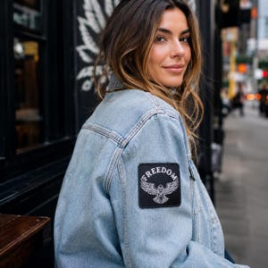 May include: A light blue denim jacket featuring a square black patch on the upper sleeve. The patch displays a white graphic of an eagle with wings spread wide and the word FREEDOM arched in white capital letters above the eagle illustration.