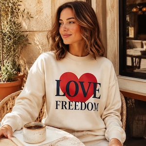 Op de afbeelding: Een crèmekleurig sweatshirt met lange mouwen en een grote rode hartafbeelding op de borst. Het woord LOVE staat in dikke donkerblauwe letters over het hart gedrukt, met het woord FREEDOM in bijpassende donkerblauwe letters eronder.