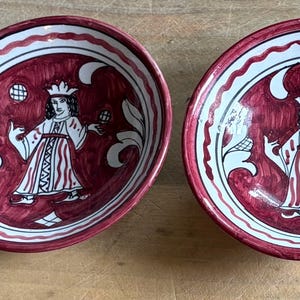 May include: Two matching ceramic bowls with a deep red interior and white and red trim. Each bowl features a central illustration of a figure in a crown and robe, holding objects. The bowls are likely for serving food.