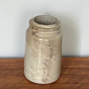May include: A cylindrical ceramic jar with a light beige, off-white glaze and a textured, weathered appearance. The jar features a prominent, raised rim around the opening and a smooth, rounded base, showcasing a mottled, aged finish throughout.