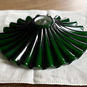 May include: A dark green glass lamp shade featuring a pleated, fan-like design with a circular opening at the center. The shade has a glossy finish and a scalloped edge, creating a radial pattern that extends from the top center to the outer rim.